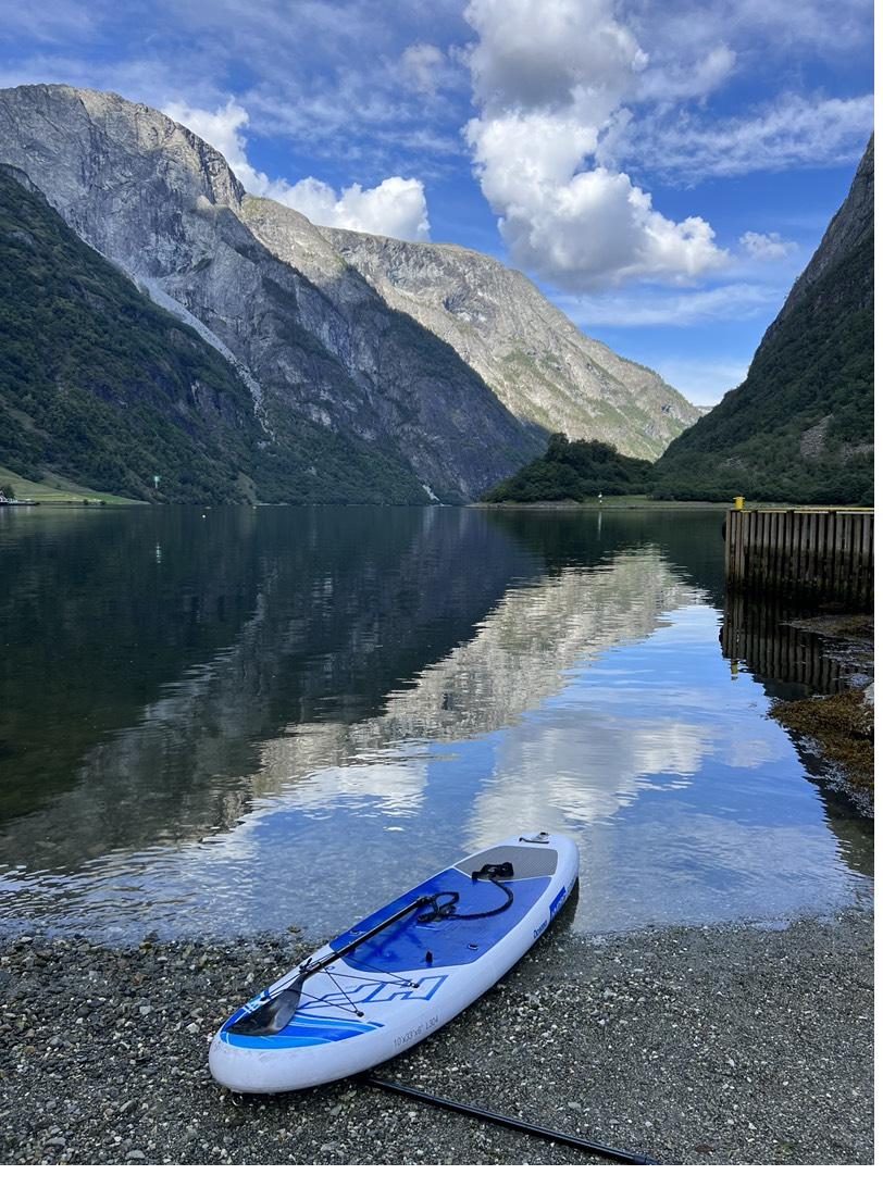 A paddleboard on a beach by a mirror-still fjord between steep mountains